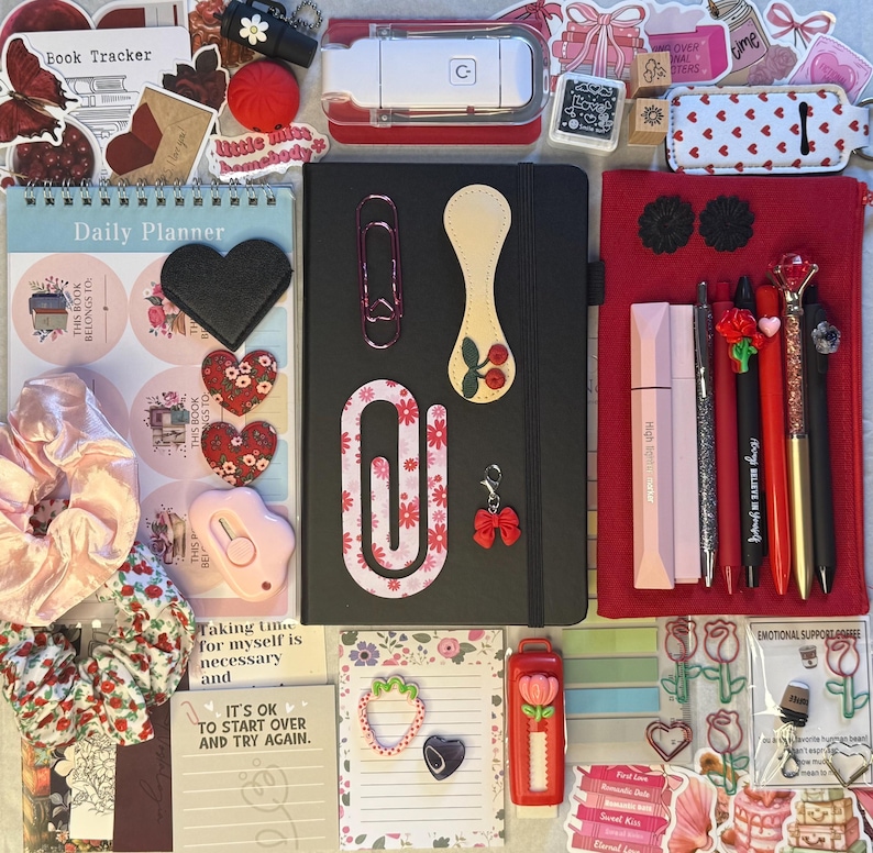 K&ouml;nnte beinhalten: Eine Sammlung von Schreibwaren, darunter ein schwarzes Notizbuch, Stifte, Aufkleber und ein Tagesplaner. Die Artikel sind in Rot-, Rosa- und Schwarzt&ouml;nen gehalten und mit Herz- und Blumendesigns versehen. Es gibt auch B&uuml;roklammern, einen Schleifenanh&auml;nger und eine rosa Haargummi.