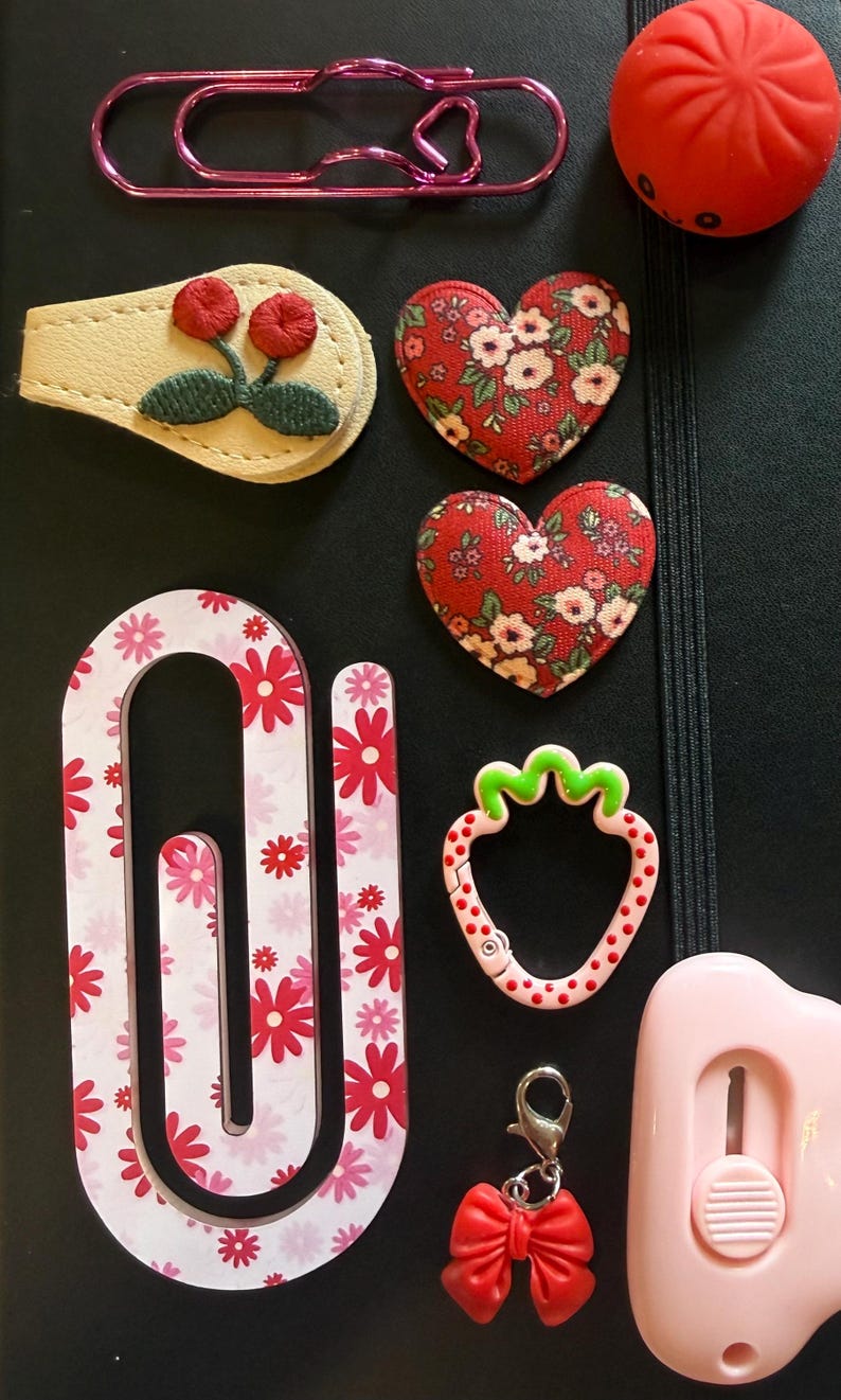 K&ouml;nnte beinhalten: Verschiedene Schreibwaren, darunter eine rosafarbene B&uuml;roklammer, ein roter herzf&ouml;rmiger Stressball, zwei herzf&ouml;rmige Stoffaufn&auml;her mit Blumenmustern und eine rosa-wei&szlig;e B&uuml;roklammer mit Blumenmuster. Enth&auml;lt auch einen Erdbeer-Charm.