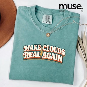 Op de afbeelding: Een turquoise Comfort Colors T-shirt met de tekst "MAKE CLOUDS REAL AGAIN" in een retro lettertype. Een zilveren ketting met een hanger ligt op het shirt. Het shirt is opgevouwen en wordt op een wit oppervlak getoond.