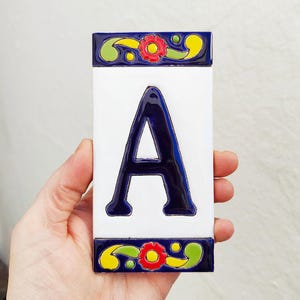 May include: A rectangular ceramic tile with a white background and a large, dark blue letter "A" in the center. The top and bottom edges feature a decorative border with red flowers, yellow accents, and green leaves. The tile is held in a hand.
