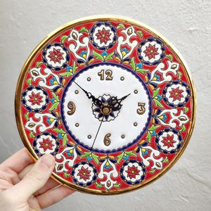 May include: A round, decorative wall clock with a gold-colored rim. The clock face is white with black hands and Arabic numerals. The outer rim features a red, blue, and green floral pattern. The clock is being held by a person.