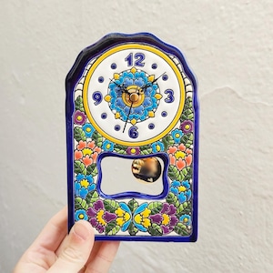 May include: A decorative ceramic clock with a blue, white, yellow, and floral design. The clock face has black hands and the numbers 12, 3, 6, and 9. The clock has a rectangular shape with a curved top and a small mirror.