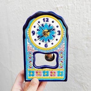 May include: A decorative ceramic clock with a blue, yellow, and white floral design. The clock face has black numbers and hands, and a pendulum. The clock is approximately 7 inches tall and 4 inches wide.