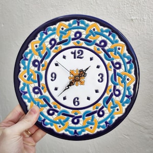 Könnte beinhalten: Runde Wanduhr aus Keramik mit weißem Zifferblatt und schwarzen Zeigern. Die Uhr hat ein dekoratives blaues, gelbes und weißes Muster am Rand. Die Zahlen 12, 3, 6 und 9 sind auf dem Zifferblatt sichtbar.