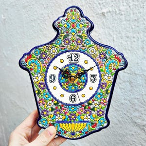 Könnte beinhalten: Eine handbemalte Keramik-Wanduhr mit einem lebendigen Blumendesign. Das Zifferblatt der Uhr ist weiß mit schwarzen Ziffern und Zeigern. Der Rand der Uhr ist tiefblau, und die Gesamtform ist verziert und dekorativ.