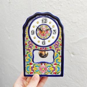 May include: A decorative ceramic clock with a white clock face featuring black numerals and hands. The clock is adorned with colorful floral patterns in pink, yellow, blue, and purple, and has a pendulum. The clock is held by a hand.