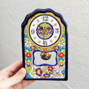 May include: A ceramic clock with a navy blue scalloped frame. The clock face is white with black numerals and gold accents. The clock is decorated with colorful floral patterns and a gold pendulum. The clock measures approximately 6 inches tall.