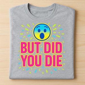 Op de afbeelding: Grijs T-shirt met ronde hals, met een verbaasd emoji en de tekst "BUT DID YOU DIE" in roze. De emoji is blauw en geel, omringd door groene en blauwe stralen.