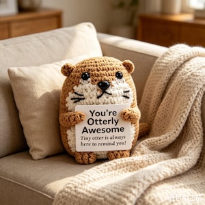 May include: A crocheted otter plushie in shades of brown and white, holding a sign that reads "You're Otterly Awesome." The plushie has small ears, a white face, and black whiskers. The text below the main message says "Tiny otter is always here to remind you!"