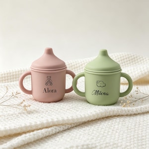 Pode incluir: Dois copos de transição em silicone com pegas e tampas. O copo rosa seco apresenta um ícone de urso e o nome Alora. O copo verde sálvia apresenta um ícone de sol e nuvem e o nome Milena. Ambos os copos têm um acabamento mate e liso.