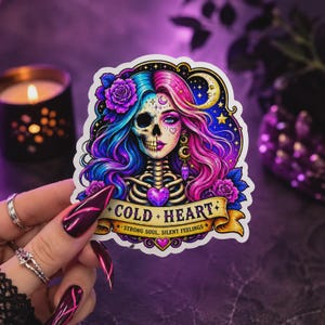 May include: A die-cut sticker featuring a split-face design of a woman and a skull with flowing blue and pink hair, purple roses, and a crescent moon. A gold banner at the bottom displays the text COLD HEART and STRONG SOUL, SILENT FEELINGS.