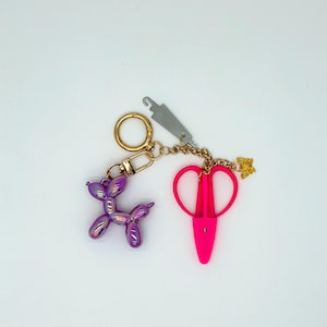 May include: A gold-toned keychain featuring a purple iridescent balloon dog charm, a bright pink pair of craft scissors, a small gold butterfly charm, and a silver-toned metal tool attachment, all connected by a gold-toned chain and ring.