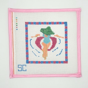 May include: A square needlepoint canvas featuring a person in a blue swimsuit and green hat floating on a pink inner tube. The design is framed by a blue and pink checkered border, finished with pink fabric binding, and includes the letters SC.