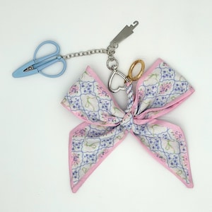 May include: A fabric bow keychain featuring a white background with a blue grid pattern, pink and blue floral motifs, and light green ribbons. It includes a silver chain, a light blue scissor-shaped charm, a metal hook, and a heart-shaped ring.