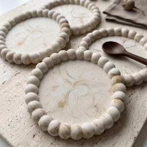 Può includere: Due sottobicchieri circolari caratterizzati da una superficie liscia color crema con vortici color marrone chiaro simili al marmo. Ogni sottobicchiere è incorniciato da un bordo di piccole perle sferiche uniformi con finitura opaca.