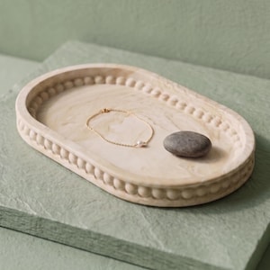 May include: An oval-shaped beige tray with a beaded rim edge. Inside the tray rests a delicate gold chain bracelet featuring a single small white pearl and a smooth, dark grey river stone. The tray has a marbled texture and light cream finish.