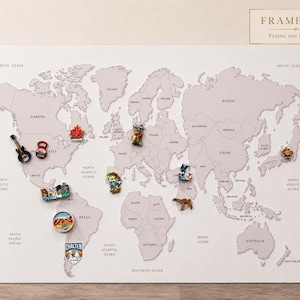 May include: A light gray world map wall art print featuring black text labels for countries and oceans. The map is decorated with various colorful souvenir magnets placed across different regions. A small box in the top right corner reads FRAMELESS.