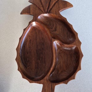 May include: A dark brown wooden serving tray shaped like a pineapple with three distinct carved compartments. The tray features a textured outer edge and a carved leaf design at the top, showcasing a natural wood grain pattern throughout the surface.