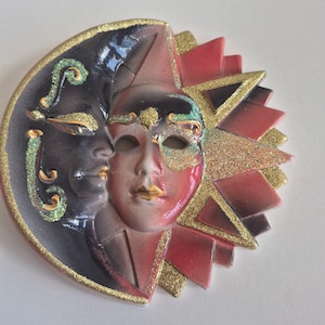 May include: A decorative wall mask featuring a dual-faced design. One side depicts a dark crescent moon with green glitter accents, while the other side shows a sun face in shades of red and gold glitter. The piece has a glossy finish.
