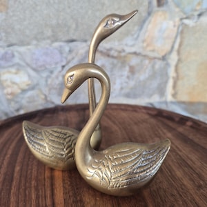 May include: A pair of brass swan figurines with polished metallic surfaces and etched wing details. One swan is positioned in the foreground with its neck curved downward, while the second swan stands behind it with its neck extended upward.