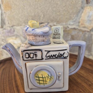 May include: A ceramic teapot shaped like a washing machine with a light blue handle and spout. The top features a laundry basket with yellow clothing. The front displays a digital timer, the text PANDA BRAND, and a circular door with a yellow grid.
