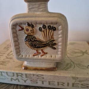 May include: A square-shaped ceramic bottle with a light beige finish. The front features a raised, embossed relief of a stylised bird with brown, black and orange accents. The bird has a fanned tail and an open beak. The bottle has a narrow neck.