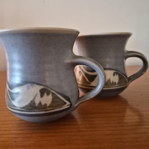 May include: Two ceramic mugs with a speckled light blue finish. Each mug features a curved handle and a decorative band near the base, displaying a stylised white and dark grey mountain landscape pattern against the blue speckled background.