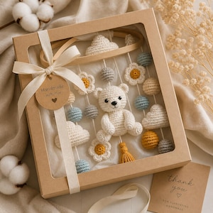 Puede incluir: Una caja de regalo cuadrada de cartón con ventana transparente que contiene un móvil de ganchillo con un oso blanco, nubes, flores y esferas en azul, gris y amarillo. Una cinta crema con una etiqueta que dice Handmade with love rodea la caja.