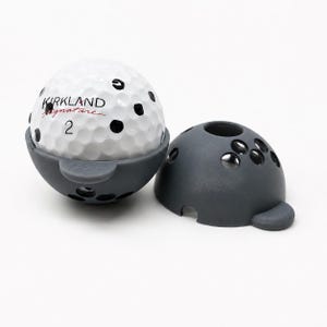 Puede incluir: Una pelota de golf blanca con el texto "KIRKLAND Signature" y el número "2" está parcialmente dentro de un soporte gris. Hay una tapa gris a juego al lado. La pelota tiene hoyuelos negros. Accesorio de golf.