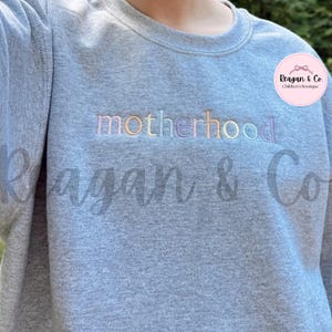 May include: A gray sweatshirt with the word "motherhood" embroidered in pastel colors across the chest. The sweatshirt also features the text "Reagan & Co" and a pink circular logo with text.