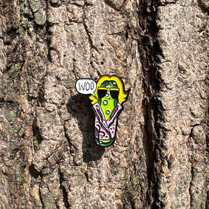 May include: A metal enamel pin shaped like a green pickle wearing black sunglasses and a purple patterned robe with yellow hair. A white speech bubble extending from the left side of the pickle contains the black text WOO in capital letters.