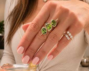 Può includere: Un anello in oro con tre pietre preziose di peridoto verde a taglio ovale disposte in fila, indossato sul dito medio. Un secondo anello in oro con diverse piccole pietre chiare a taglio rotondo e baguette è indossato sull'anulare.