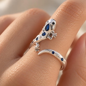 Könnte beinhalten: Ein silberfarbener, offener Ring in Form einer Eidechse, die sich um den Finger windet. Die Eidechse hat eine glatte, metallische Oberfläche mit kleinen, dunkelblauen, tropfenförmigen und runden Edelsteinakzenten auf dem Rücken und Schwanz.