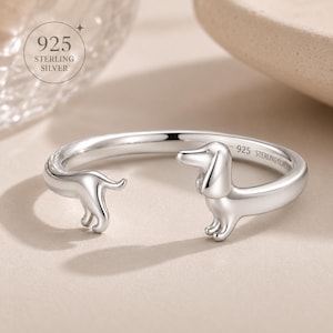 Puede incluir: Un anillo abierto de plata de ley 925 pulida con un diseño de perro salchicha. La banda del anillo forma el cuerpo del perro, con la cabeza y las patas delanteras en un extremo y las patas traseras y la cola en el otro. Incluye la inscripción 925 STERLING SILVER.