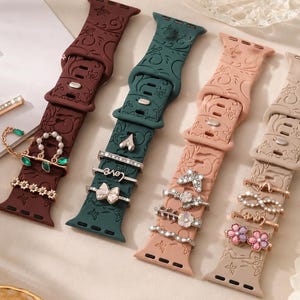 Puede incluir: Cuatro correas de reloj coloridas con dijes decorativos. Las correas son en tonos burdeos, verde azulado, rosa rubor y beige. Cada correa tiene un conjunto diferente de dijes, incluyendo lazos, corazones y diseños florales.