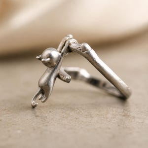 May include: A silver ring featuring a small cat clinging to a branch. The ring has a detailed design, with the cat's body and tail curved around the branch. The ring is made of a shiny, metallic material.