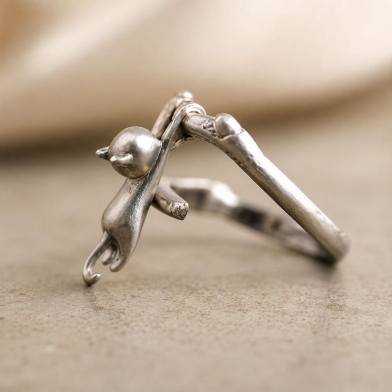 May include: A silver ring featuring a small cat clinging to a branch. The ring has a detailed design, with the cat's body and tail curved around the branch. The ring is made of a shiny, metallic material.