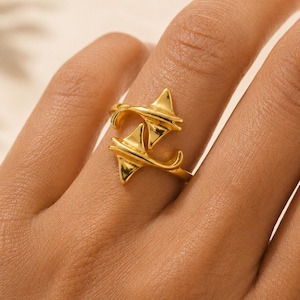 Puede incluir: Un anillo de tono dorado que presenta dos formas estilizadas de raya que se superponen en la banda. El metal tiene una superficie pulida y reflectante, y el diseño representa los cuerpos triangulares distintivos y las colas curvas de las criaturas.