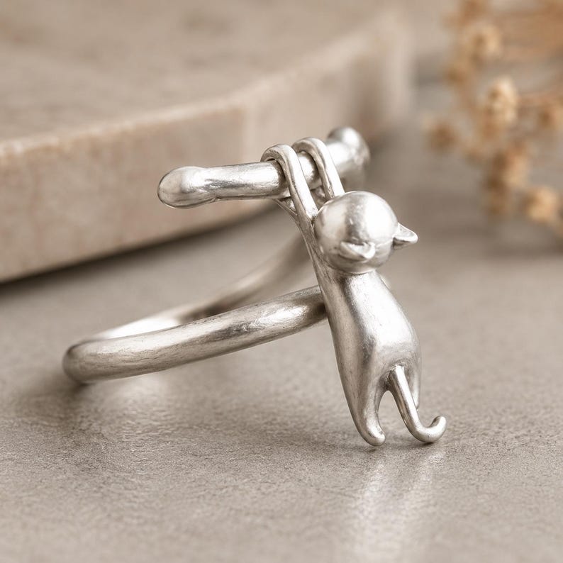 May include: A silver ring featuring a stylized cat hanging from a bar. The cat is detailed with a small head, pointed ears, and a curved tail. The ring band is smooth and simple, creating a minimalist design.