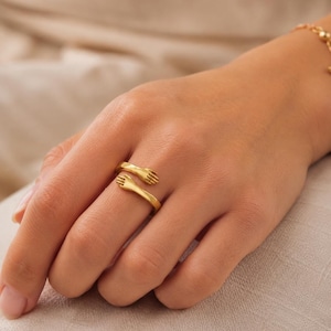 Puede incluir: Un anillo de metal en tono dorado con un diseño de dos manos que se envuelven alrededor del dedo en un movimiento de abrazo. La banda está abierta en la parte delantera, con las dos manos encontrándose sin tocarse, creando una superficie lisa.