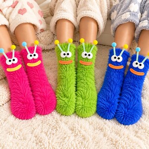 May include: Three pairs of fuzzy crew socks in bright pink, lime green, and royal blue. Each sock features a cartoon monster face with two large white eyes, an orange mouth, and two antennae topped with small blue or yellow pom-pom spheres.