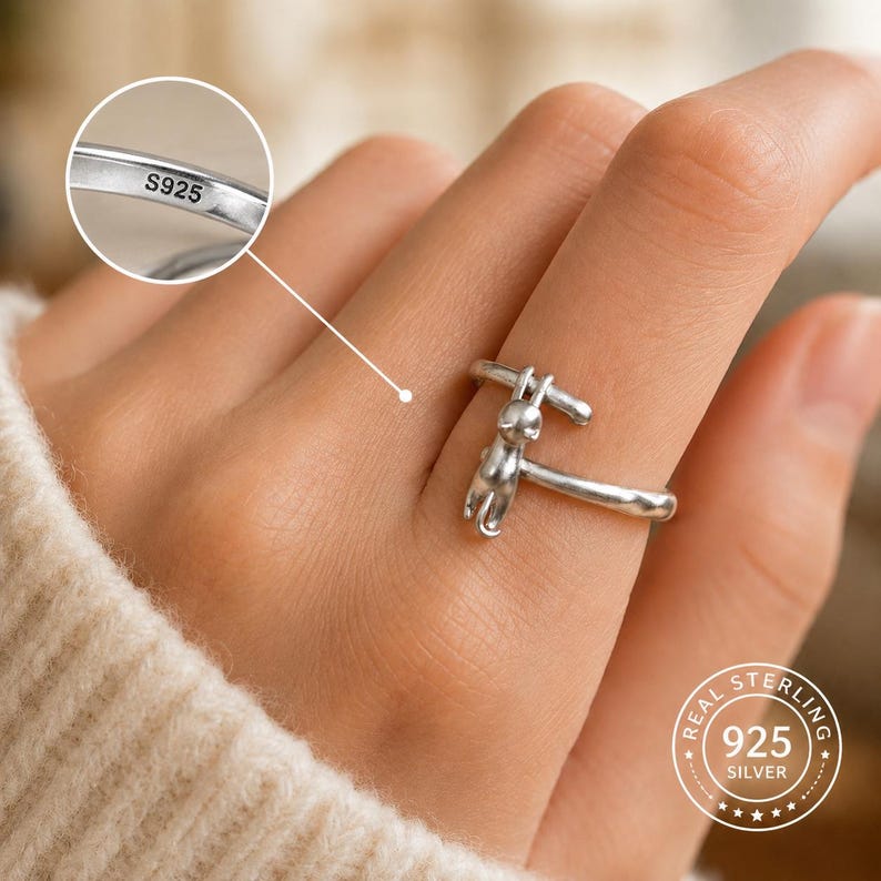 May include: A sterling silver ring featuring a cat design. The ring is stamped with "S925" and "REAL STERLING 925 SILVER". The cat is depicted in a stylized form, with a simple, elegant design. The ring is worn on a finger.
