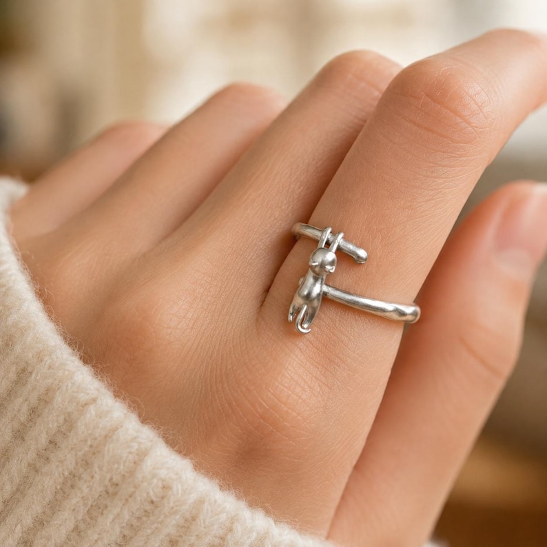 May include: A silver-colored ring featuring a small, stylized bunny design. The bunny is suspended from a bar that arches over the finger, creating a unique and whimsical look. The ring is worn on a finger.