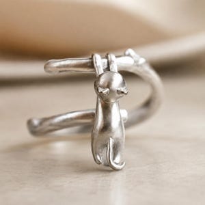 May include: A silver-colored cat ear cuff. The cuff features a stylized cat hanging from a curved bar. The cat has a smooth, rounded body and a small tail. The design is simple and elegant, suitable for cat lovers.