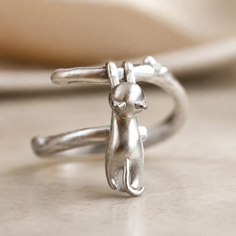 May include: A silver-colored cat ear cuff. The cuff features a stylized cat hanging from a curved bar. The cat has a smooth, rounded body and a small tail. The design is simple and elegant, suitable for cat lovers.