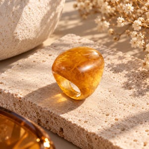 Puede incluir: Un anillo grueso y translúcido de color ámbar con una superficie lisa y redondeada, que presenta un patrón jaspeado y arremolinado en todo el material. El anillo tiene un diseño de banda gruesa y sólida con un acabado pulido.