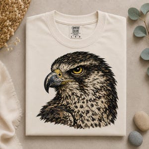 Puede incluir: Una camiseta de manga corta color crema con una ilustración detallada de una cabeza de halcón de perfil. El ave tiene plumas de color marrón oscuro y negro, un pico afilado y curvado y un ojo amarillo penetrante. Etiqueta Comfort Colors visible.