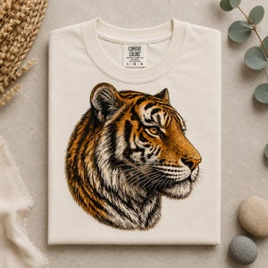 Puede incluir: Una camiseta de color crema con una ilustración detallada de una cabeza de tigre de perfil. El tigre tiene pelaje naranja, negro y blanco con ojos color ámbar penetrantes. La camiseta tiene cuello redondo y una etiqueta de la marca Comfort Colors.