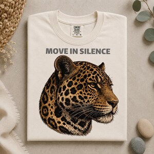 Puede incluir: Una camiseta de color crema con una ilustración detallada de una cabeza de leopardo de perfil. Sobre el leopardo, el texto MOVE IN SILENCE está impreso en una fuente sans-serif de color gris oscuro. La camiseta tiene cuello redondo y una etiqueta Comfort Colors.