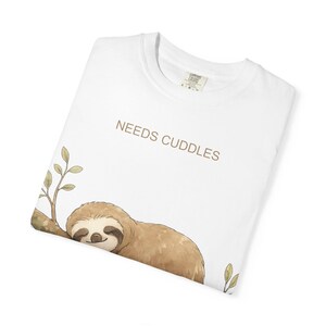 May include: White t-shirt featuring a sleeping sloth illustration and the text "NEEDS CUDDLES" in brown. The sloth is brown and tan, with a smiling face. The shirt has a crew neck and a small tag.